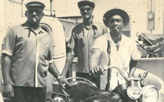 Sepia-toned photograph of three Black men posing with a motorcycle.
